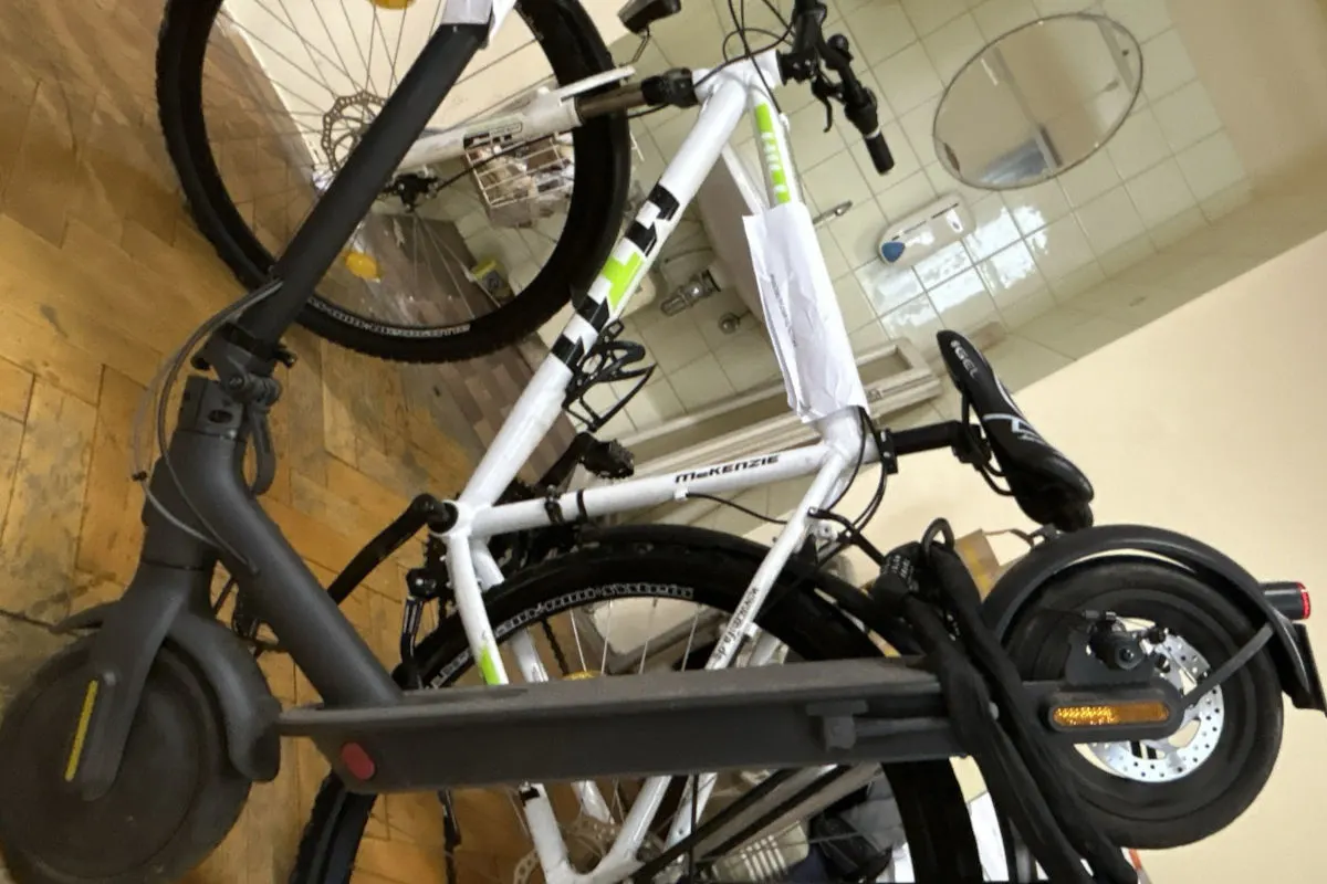In einem Raum mit gefliestem Waschbeckenbereich stehen ein weißes Fahrrad der Marke McKenzie und ein schwarzer E-Scooter nebeneinander auf Holzboden.