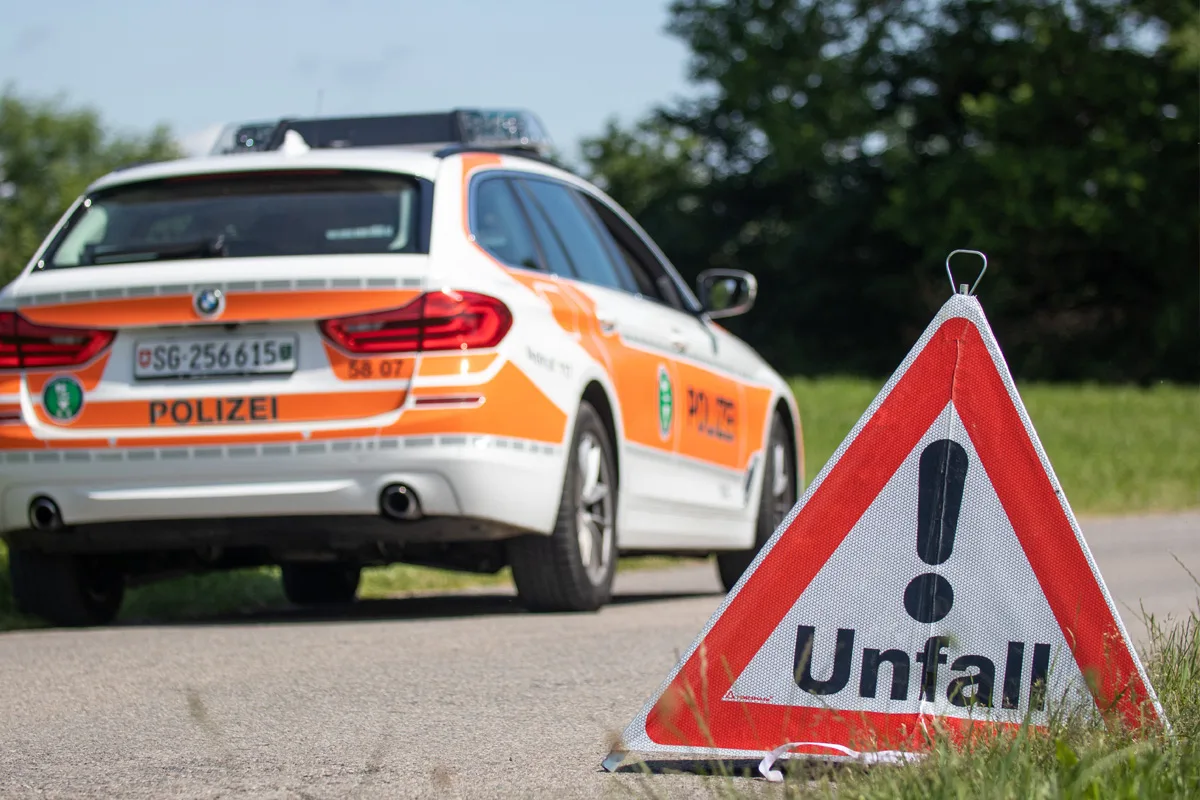Ein weißes Polizeiauto mit orangefarbenen Streifen steht hinter einem aufgestellten Unfall-Warnschild auf einer asphaltierten Straße.