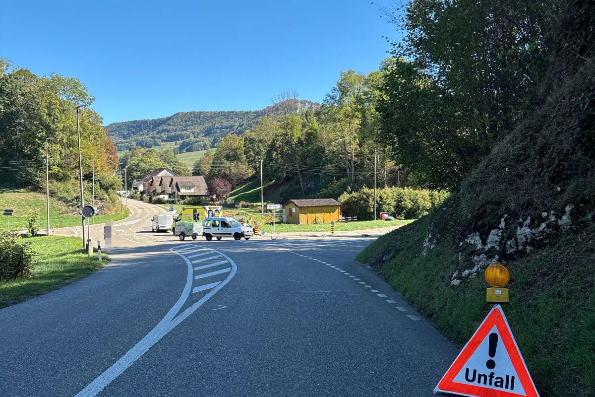 Ein Unfallwarnschild steht am Rand einer Landstraße. Im Hintergrund sind Einsatzfahrzeuge und eine ländliche Umgebung zu sehen.