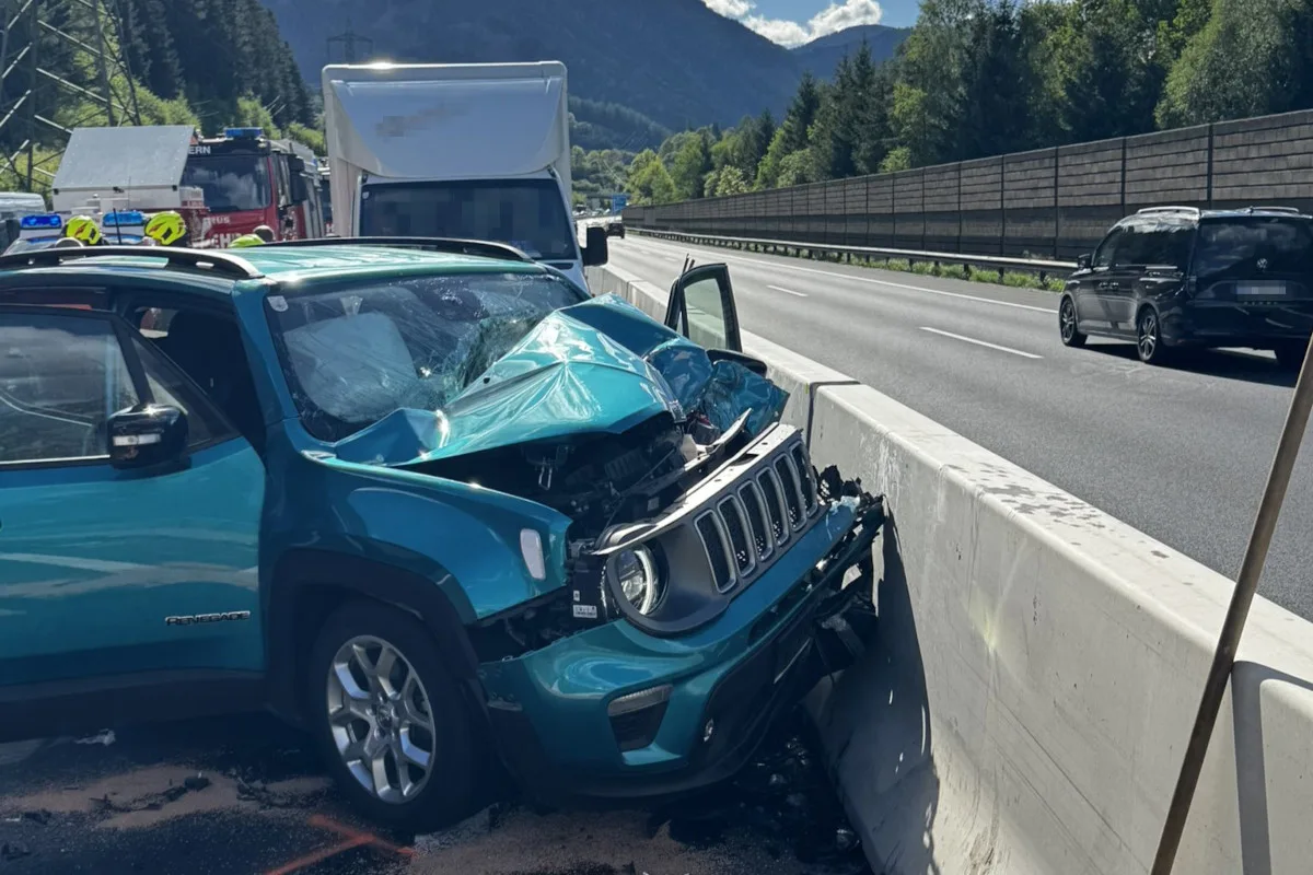 Ein blaues Auto mit erheblichem Frontschaden steht an einer Betonbarriere auf einer Autobahn. Im Hintergrund sind weitere Fahrzeuge sichtbar.
