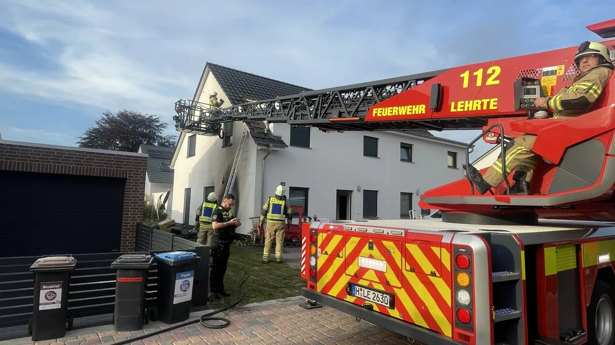Ein Feuerwehrfahrzeug mit Drehleiter ist vor einem modernen Wohnhaus im Einsatz. Mehrere Feuerwehrleute und ein Polizist stehen vor Ort.