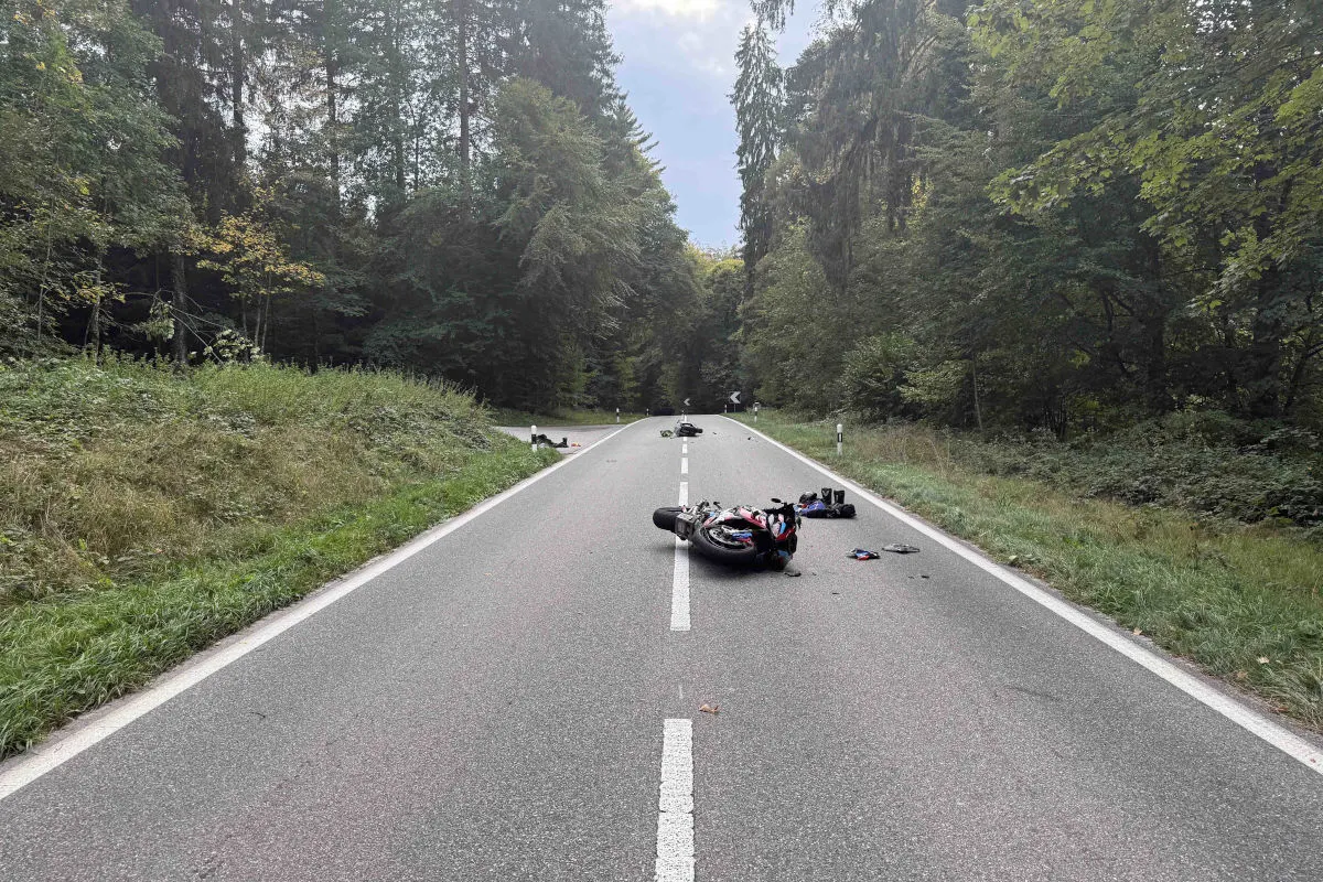 Ein umgestürztes Motorrad liegt auf der Fahrbahn einer von Wald gesäumten Landstraße, umgeben von verstreuten persönlichen Gegenständen.