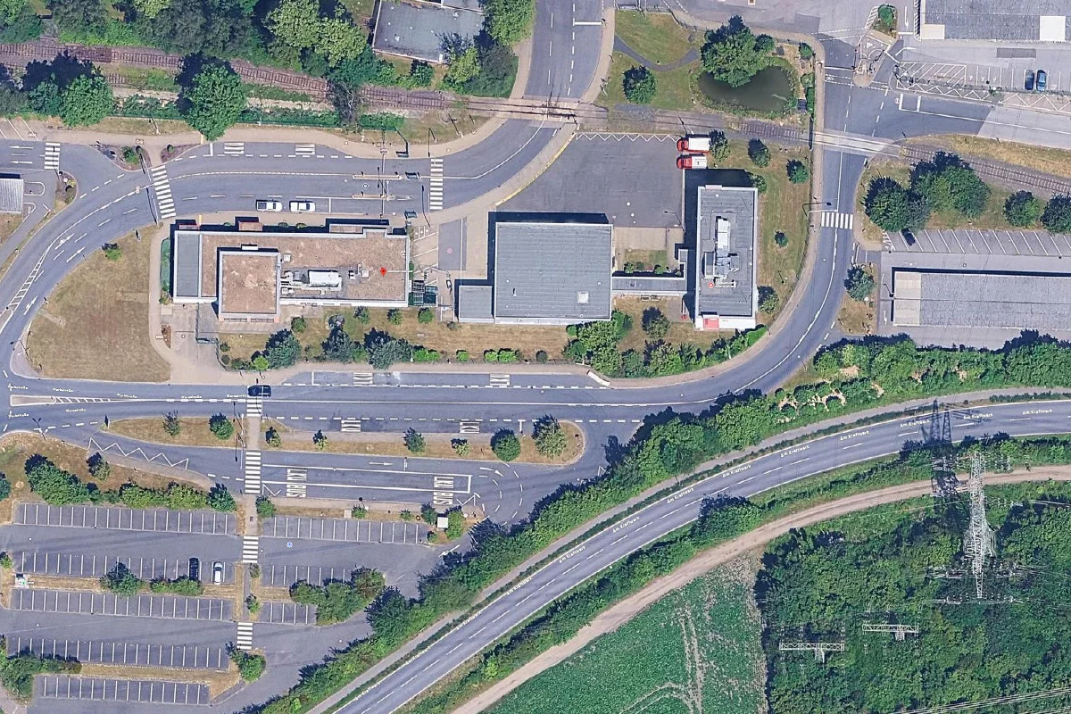 Das Bild zeigt eine Luftaufnahme einer Straßeneinmündung umgeben von Parkplätzen und Gebäuden. Zwei rote Fahrzeuge sind auf einem Parkplatz sichtbar.