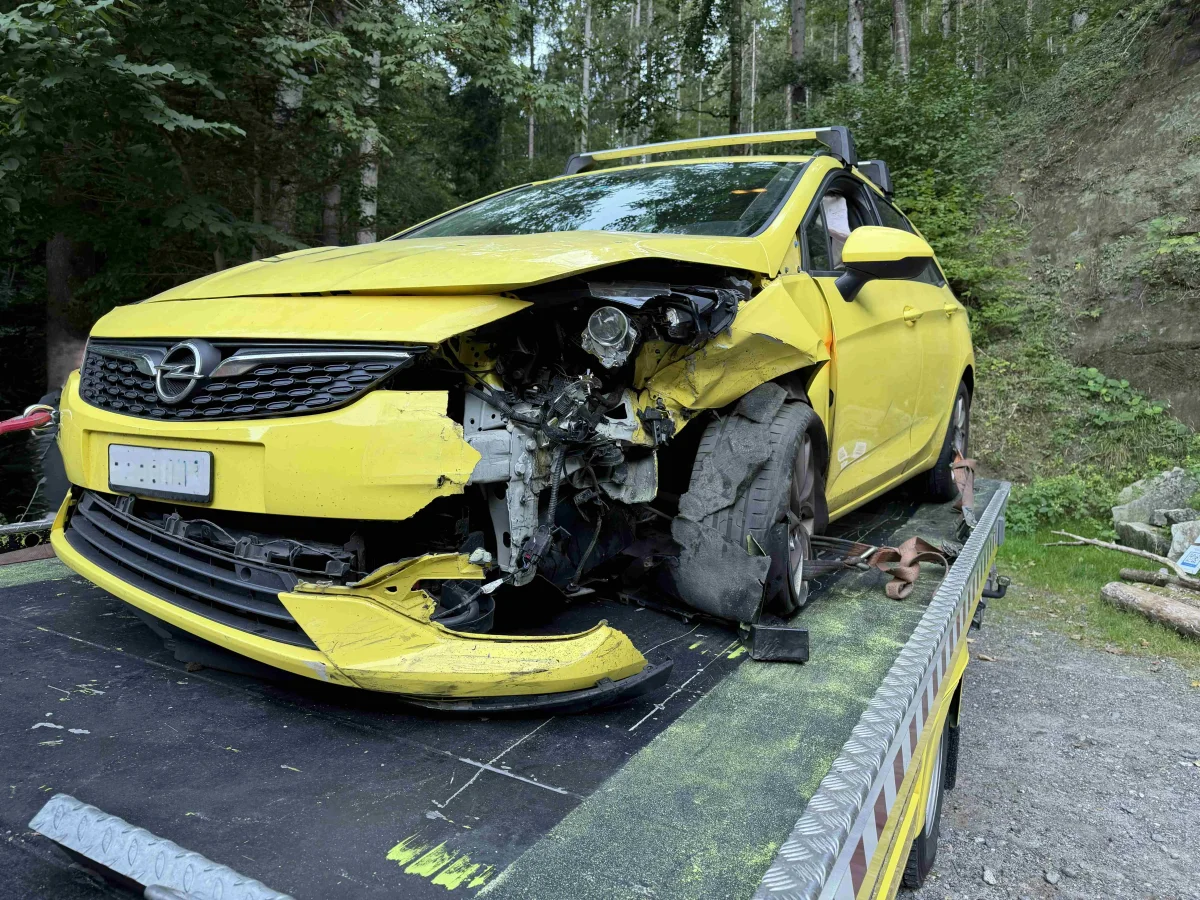 Ein gelbes Auto mit schwerem Frontschaden steht auf einem Abschleppwagen im Wald. Die linke Seite ist stark eingedrückt.