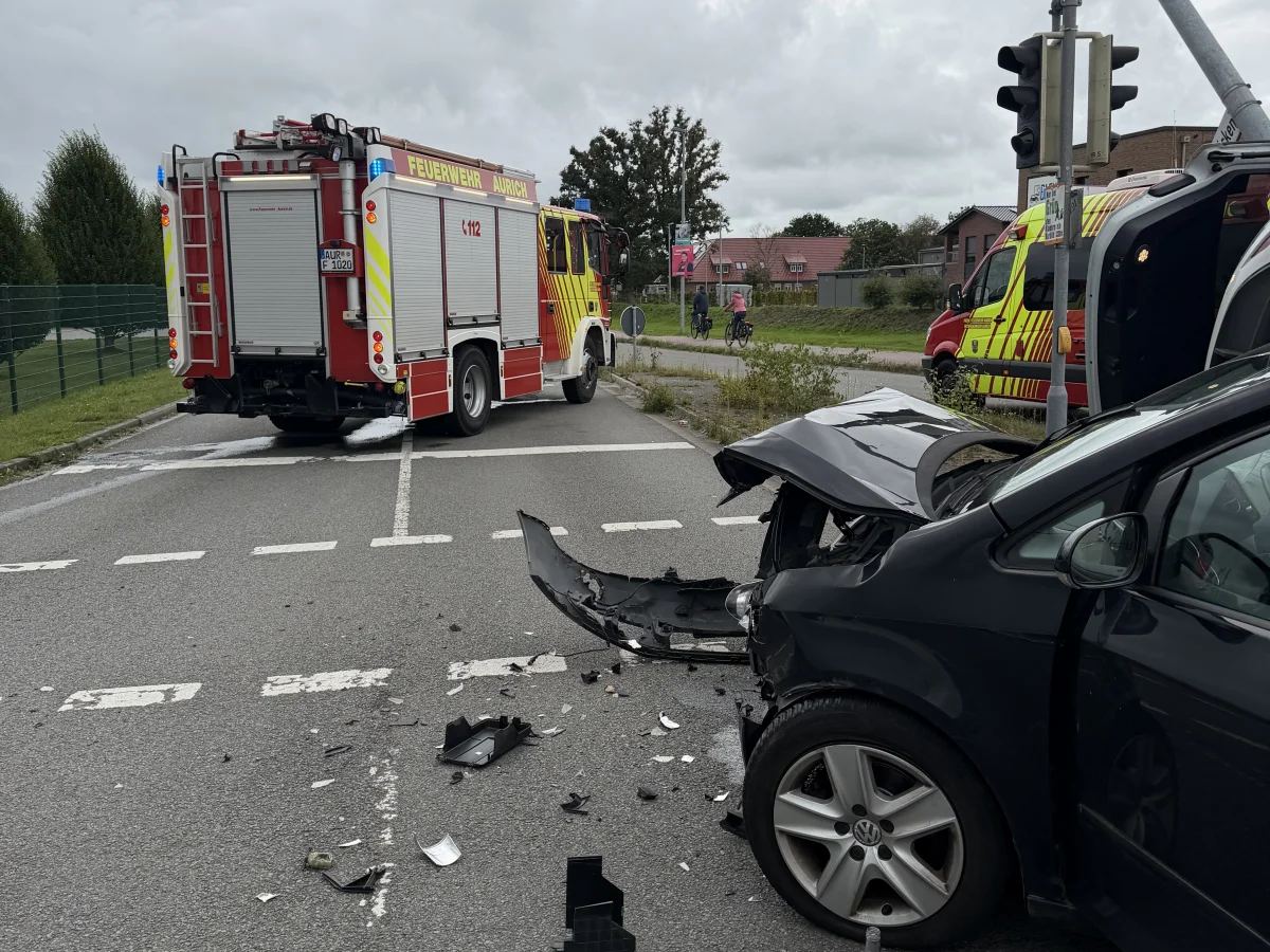 Ein schwarzer VW mit erheblichem Frontschaden steht an einer Kreuzung neben einem umgeknickten Ampelmast. Im Hintergrund sind zwei Feuerwehrfahrzeuge zu sehen.
