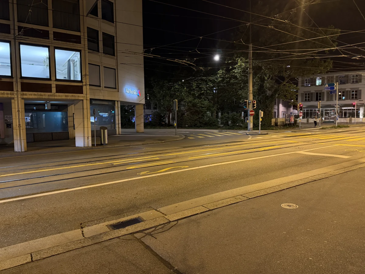 Eine nächtliche Straßenkreuzung in der Stadt ohne Verkehr, mit beleuchteten Gebäuden im Hintergrund und Straßenbahnlinien im Vordergrund.