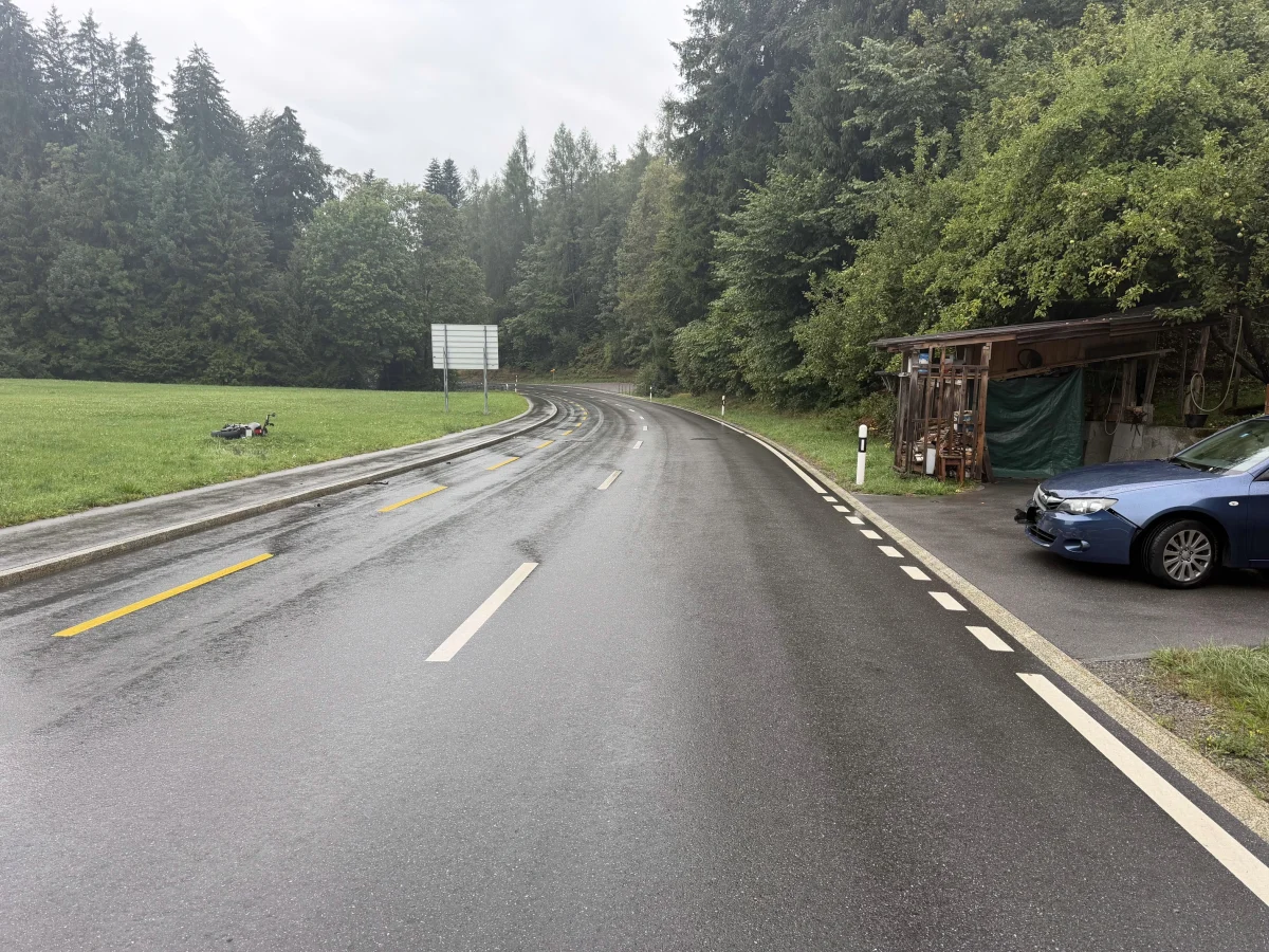 Auf einer kurvigen, regennassen Straße im Wald liegt ein Motorrad im Gras. Rechts ist ein geparktes blaues Auto vor einer Holzstruktur sichtbar.
