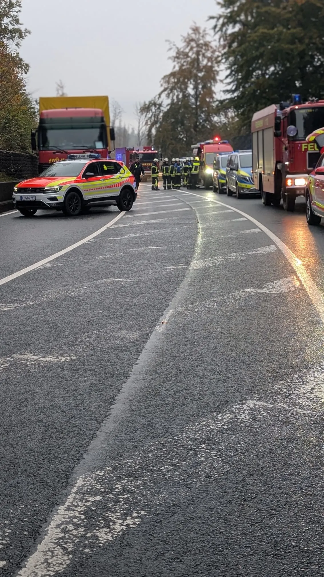 Mehrere Feuerwehr- und Rettungsfahrzeuge stehen auf einer regennassen Straße. Einsatzkräfte in Uniform arbeiten vor Ort.
