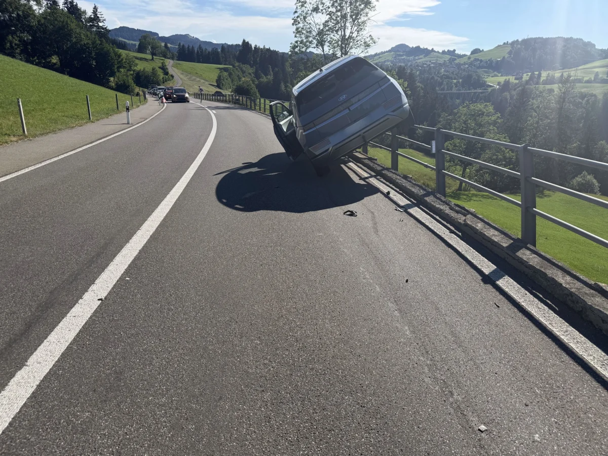 Ein silbernes Auto ist von der Fahrbahn einer kurvigen Landstraße abgerutscht und hängt am Geländer. Im Hintergrund erstreckt sich eine hügelige Landschaft unter klarem Himmel.