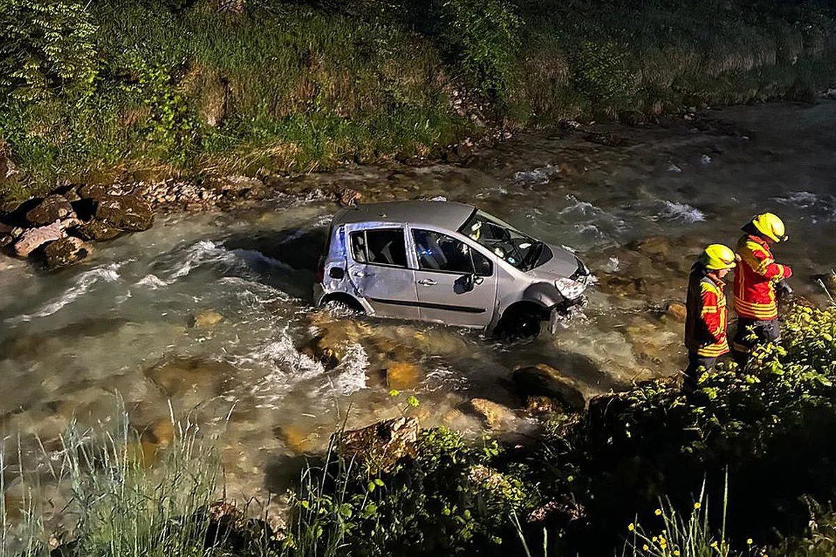 Ein silbernes Auto steht im Bachbett, während zwei Feuerwehrleute in leuchtend roten Uniformen am Ufer stehen. Es ist Nacht.