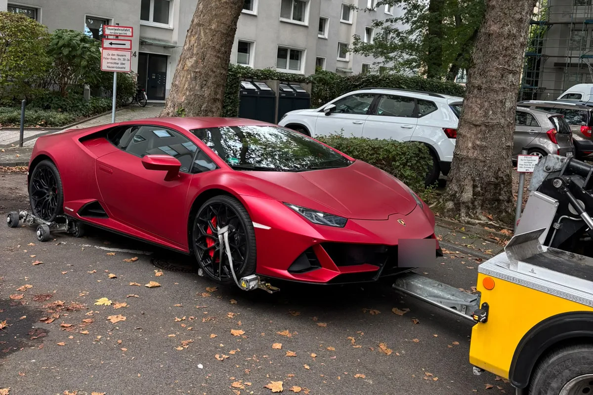 Ein roter Sportwagen steht auf einer Straße und wird von einem gelben Abschleppfahrzeug angehoben. Im Hintergrund sind Wohnhäuser und andere parkende Autos zu sehen.