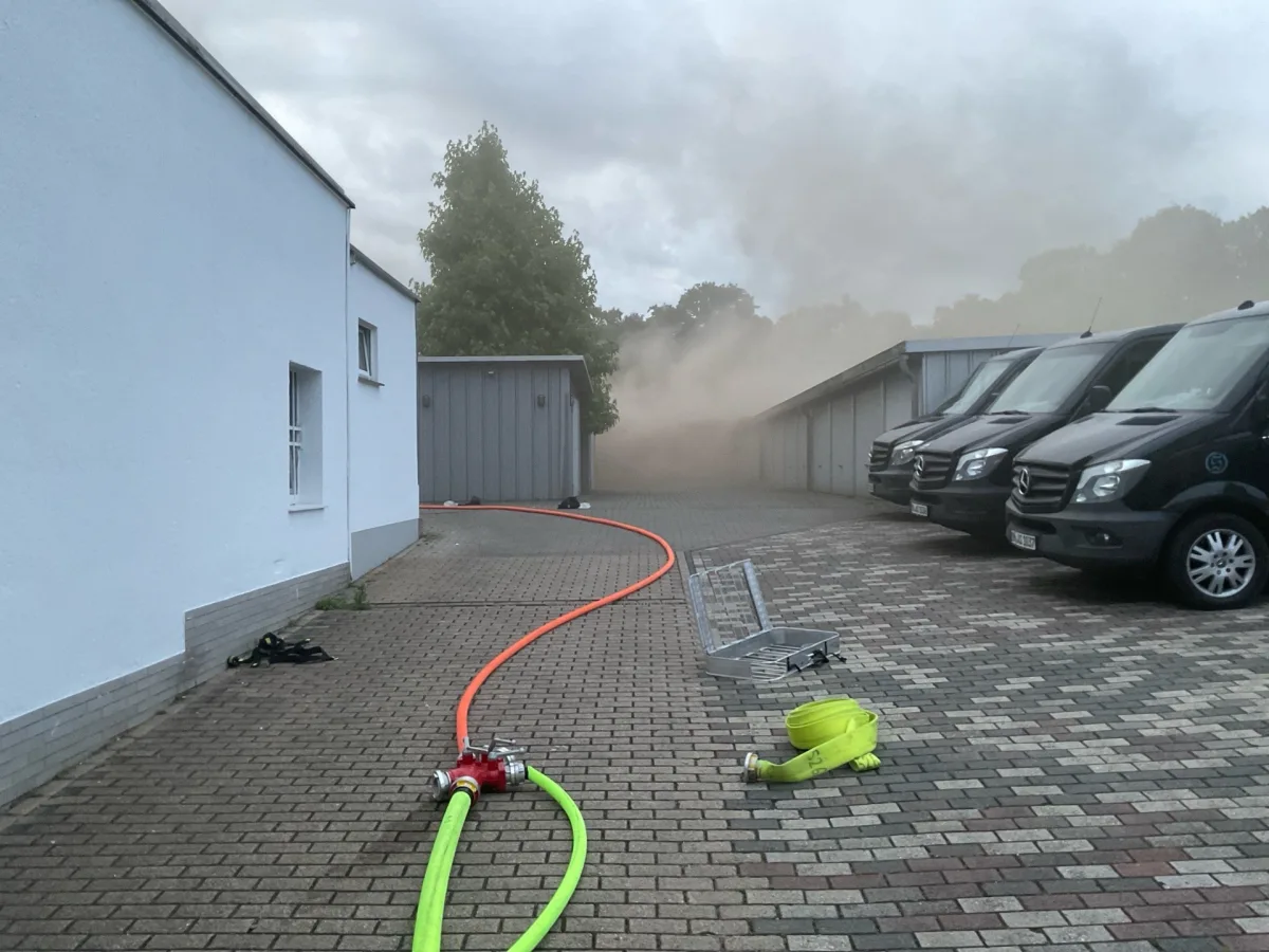 Mehrere schwarze Vans stehen in einem Hof, im Hintergrund ist Rauch zu sehen, und Feuerwehrschläuche liegen am Boden.