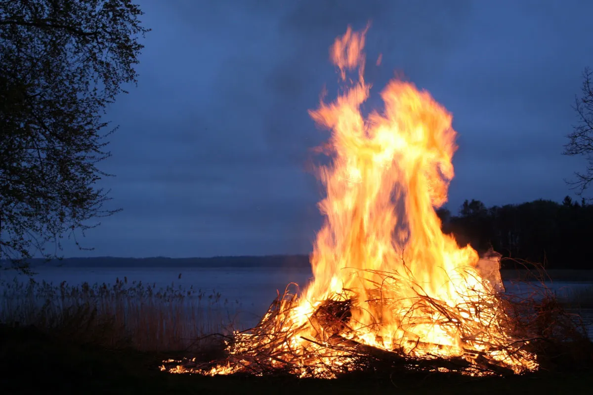 Ein großes, helles Feuer lodert am Ufer eines Sees in der Nacht. Dunkle Silhouetten von Bäumen sind erkennbar.