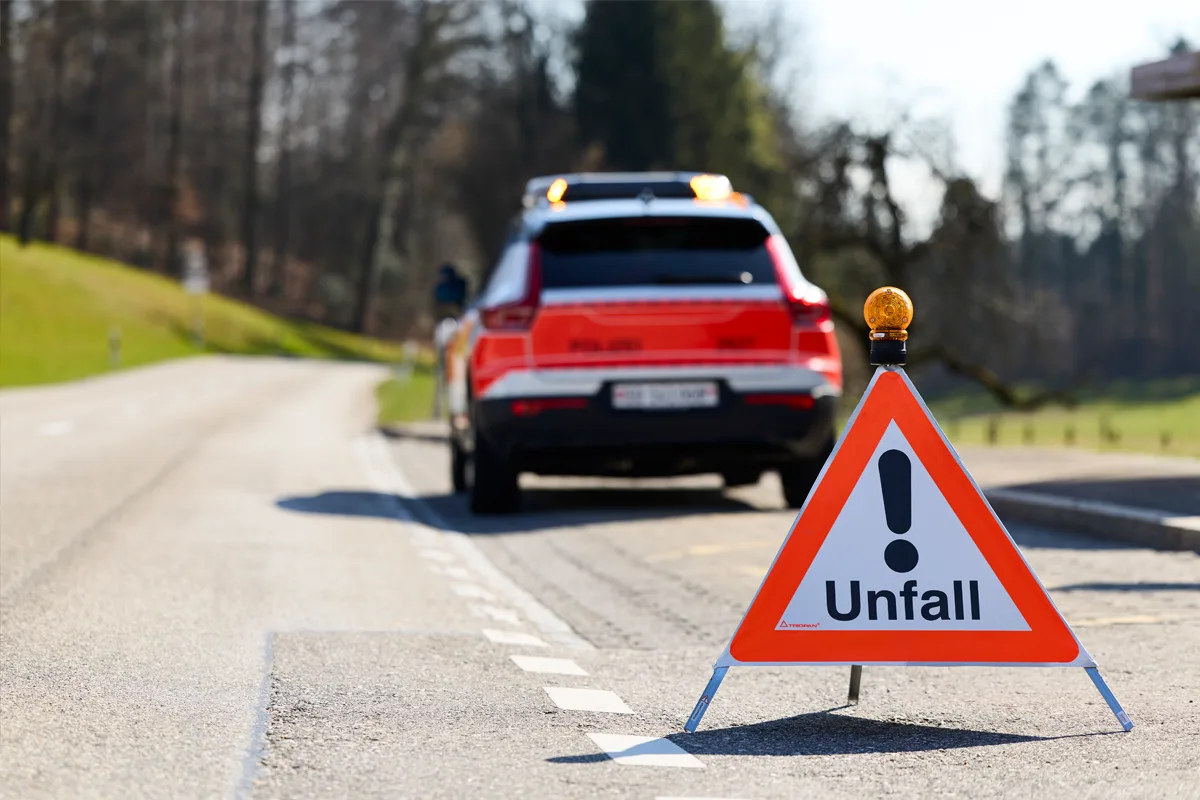 Ein Warnschild mit der Aufschrift 'Unfall' steht auf einer Straße, ein Polizeiauto ist im Hintergrund zu sehen.