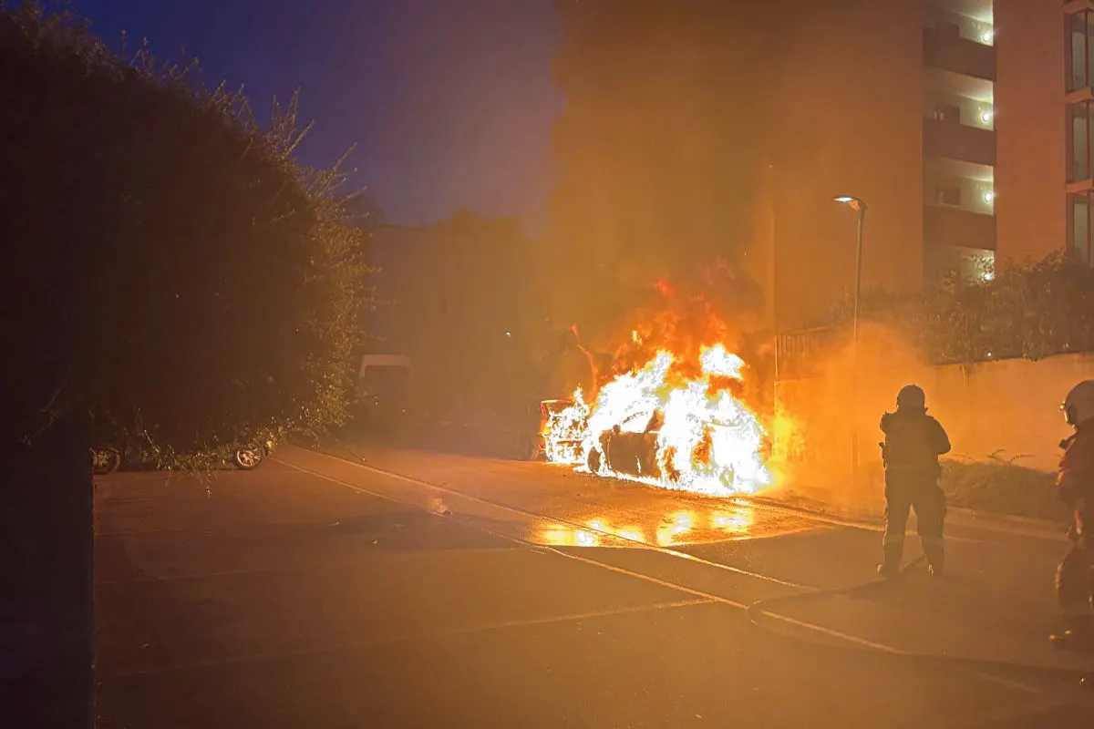 Ein Fahrzeug steht in Flammen am Straßenrand bei Nacht, während Feuerwehrleute in Schutzkleidung das Feuer überwachen.