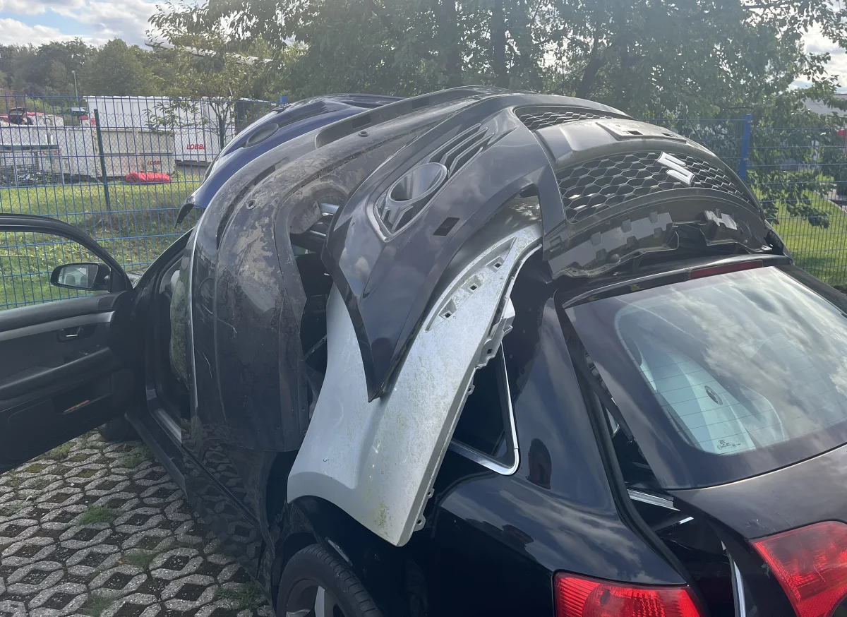 Ein schwarzes Auto steht im Freien, mit mehreren Stoßstangen obenauf, umgeben von einem Zaun und Bäumen.