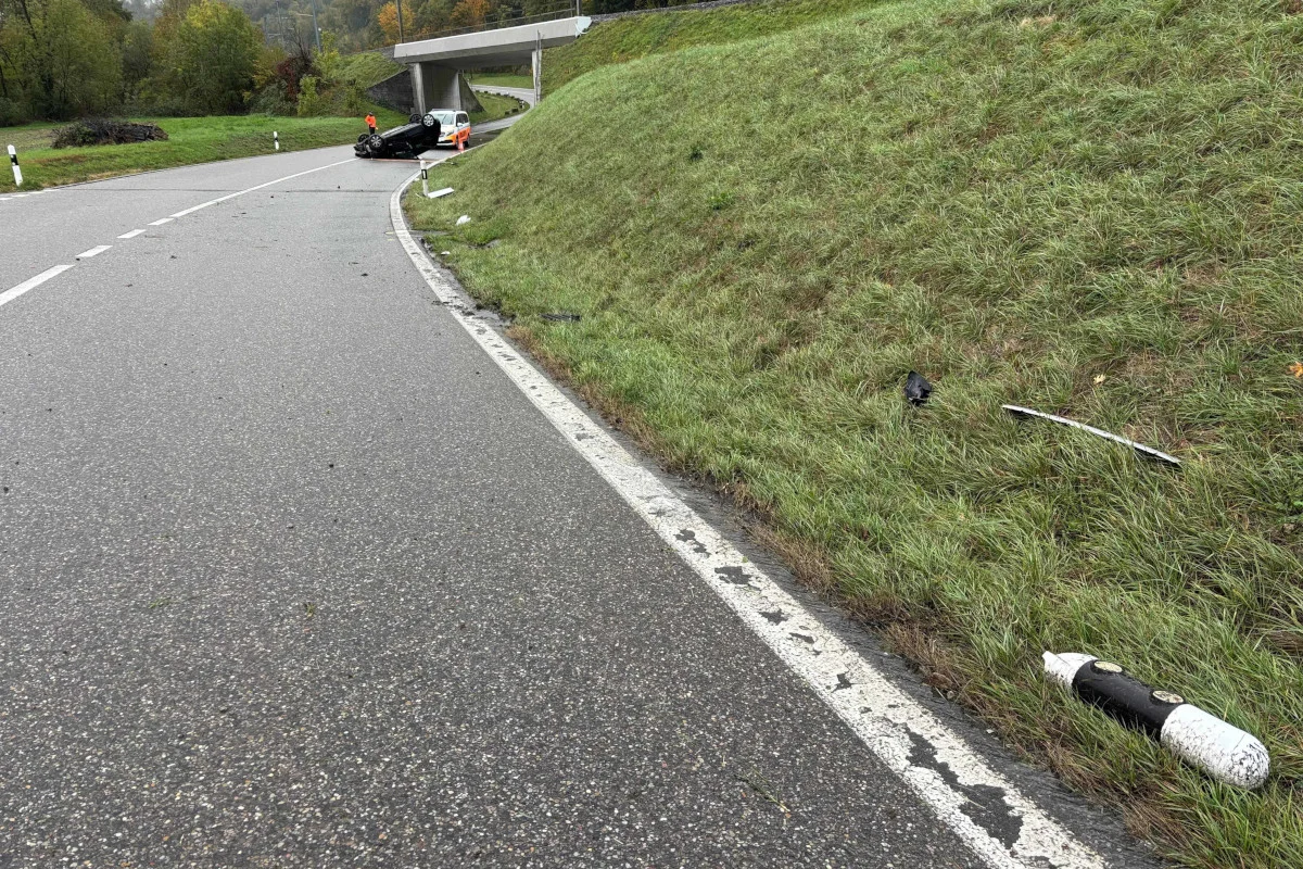 Ein Fahrzeug liegt auf dem Dach neben einer Landstraße. Trümmerteile sind über die Fahrbahn und den angrenzenden Grünstreifen verstreut, nahe einer Autobahnüberführung.