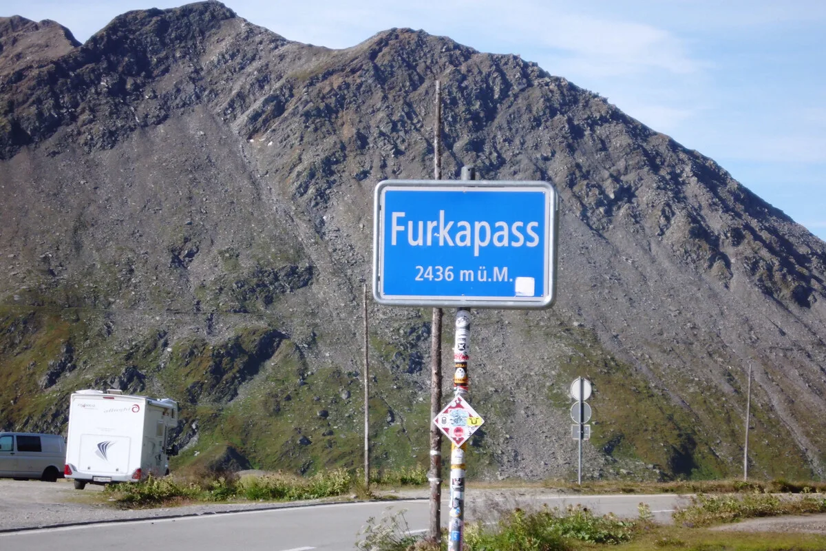 Ein blaues Schild mit der Aufschrift 'Furkapass 2436 m ü. M.' steht am Straßenrand mit einem Berg im Hintergrund.