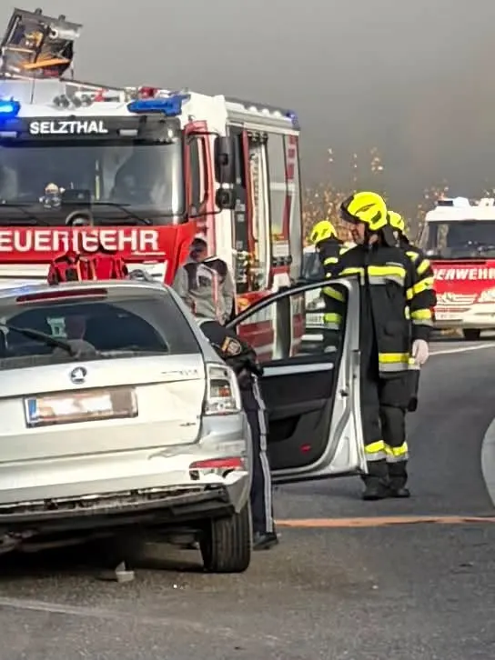 Ein Feuerwehrauto steht hinter einem beschädigten Auto, umgeben von Feuerwehrleuten in Schutzkleidung. Einsatzkräfte untersuchen das Fahrzeug.