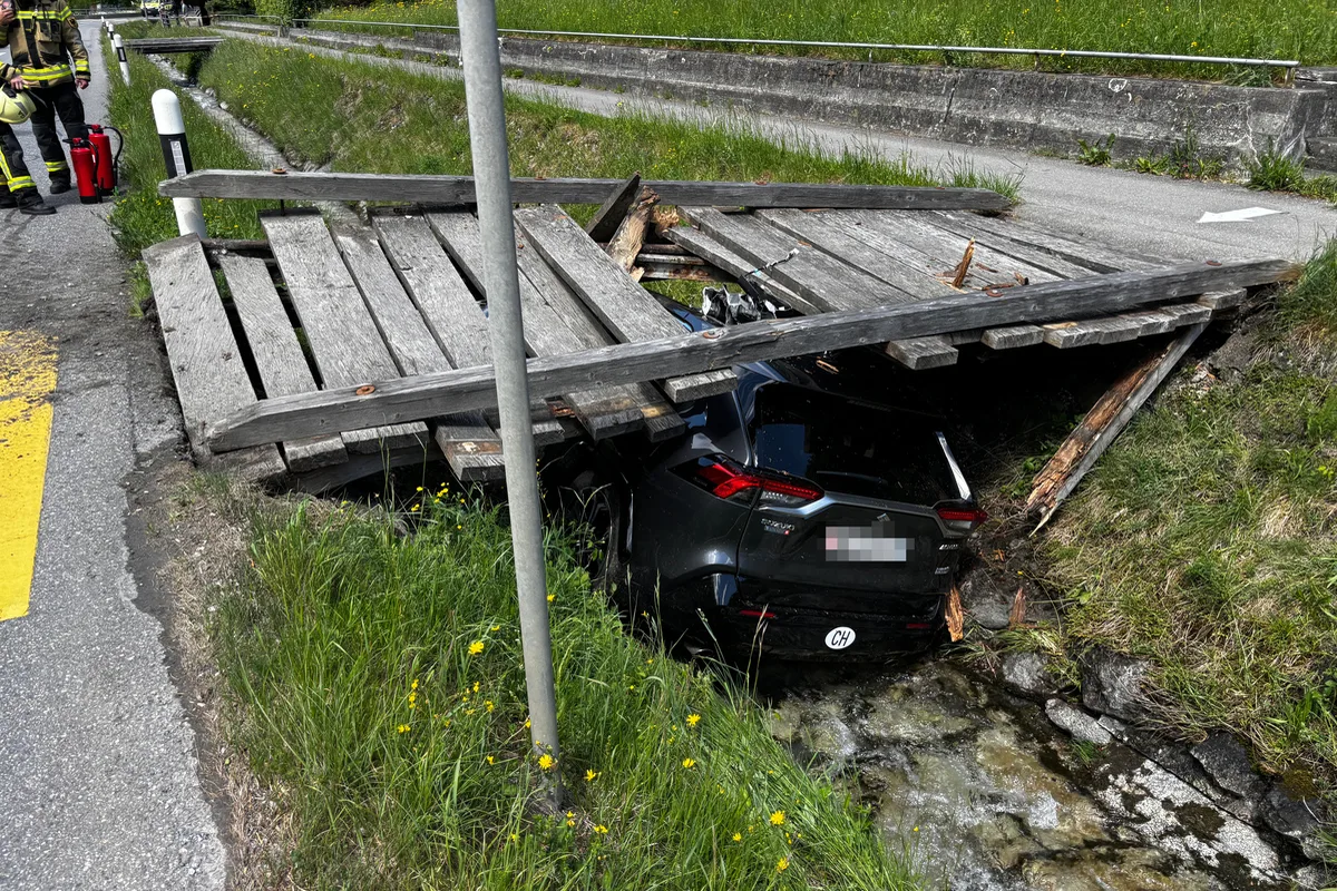 Ein schwarzes Auto ist unter einer eingestürzten Holzbrücke in einem Graben zu sehen. Ein Feuerwehrmann steht mit Feuerlöscher daneben.
