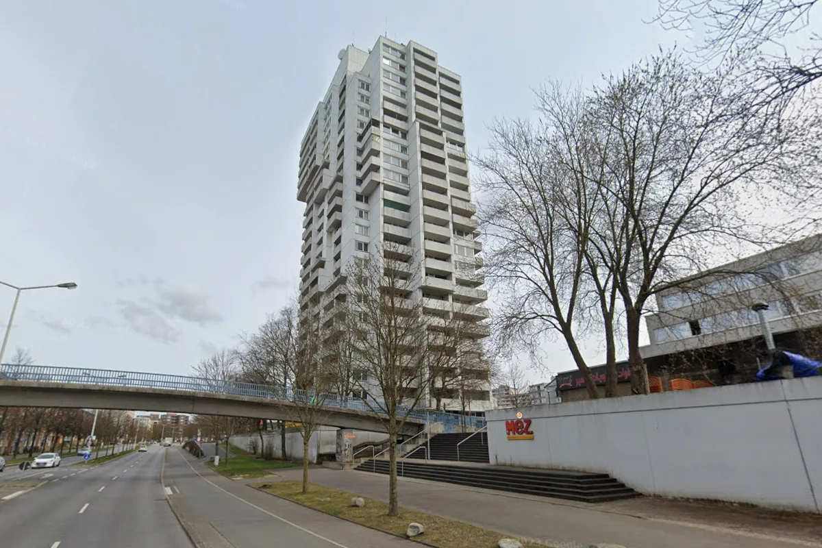 Ein modernes Hochhaus steht neben einer Straße, über die eine Fußgängerbrücke führt. Bäume ohne Laub säumen den Weg.