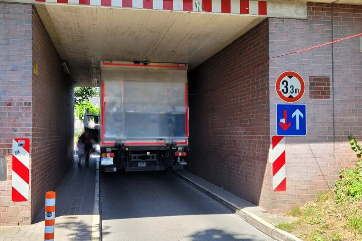 Ein großer Lastwagen steckt unter einer niedrigen Brücke fest, die Höhe ist mit 3,3 m ausgeschildert. Die Brücke verläuft über eine schmale Fahrbahn.