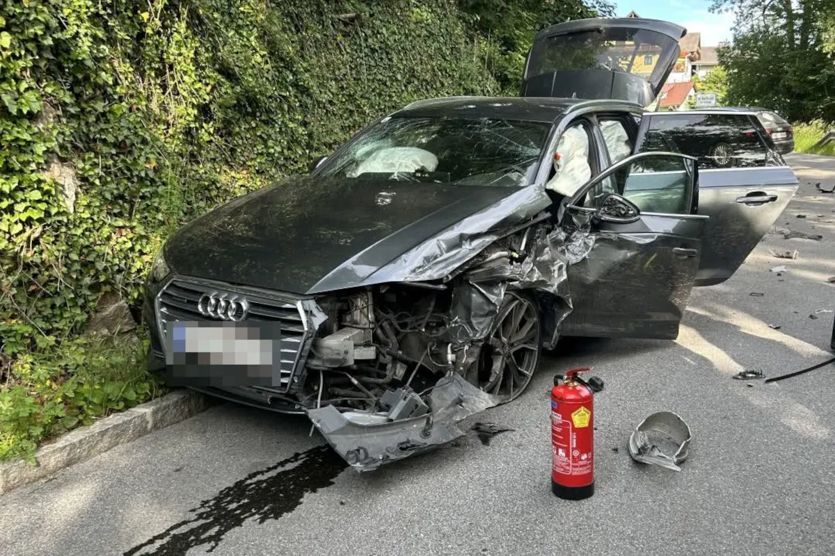 Ein schwarzer Audi steht mit stark beschädigter Frontpartie auf einer Straßenrand. Die Airbags sind ausgelöst und der Kofferraum ist offen.