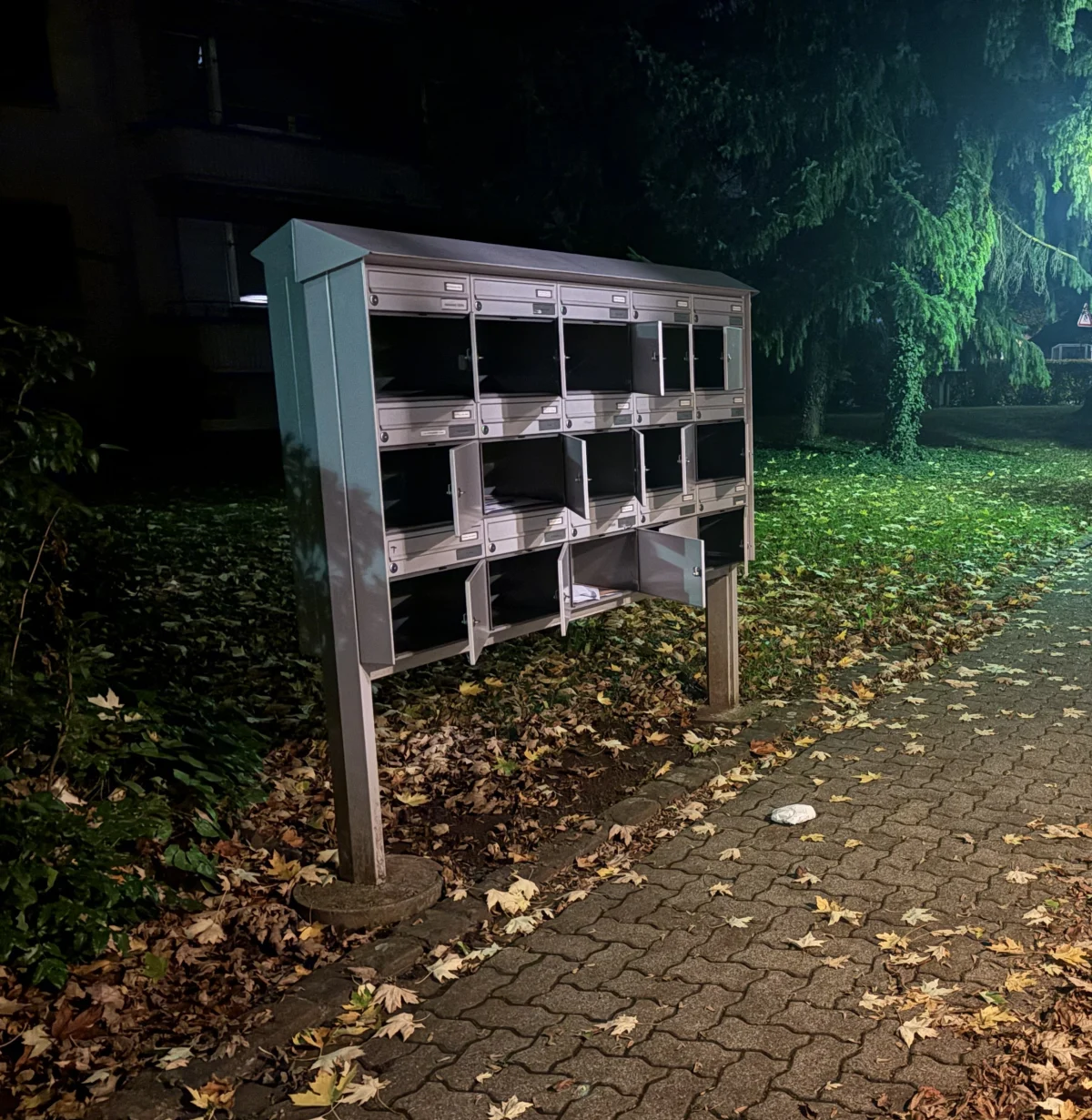 Ein großer Briefkastenständer mit mehreren offenen Fächern steht auf einem Gehweg, umgeben von herabgefallenem Laub bei nächtlicher Beleuchtung.