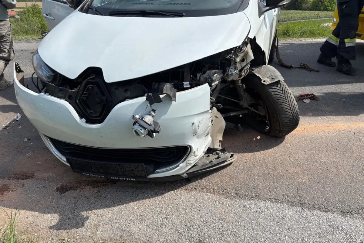 Ein weißes Auto mit schwerem Frontschaden steht still auf einer Straße. Einzelne Personen sind teilweise sichtbar, aber ihre Gesichter sind außerhalb des Bildes.