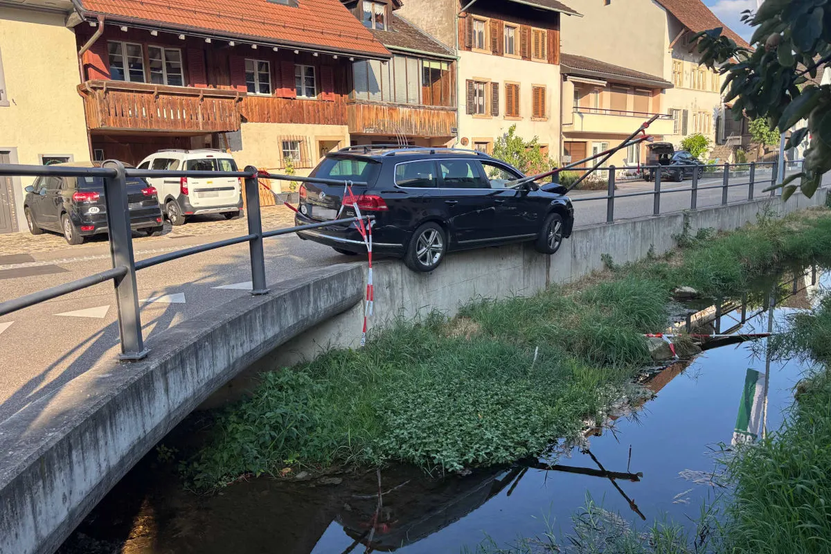 Ein schwarzer Pkw ist über die Betonbegrenzung einer kleinen Brücke gefahren und hängt teilweise in der Luft über einem Bach. Keine Personen sichtbar, die Umgebung zeigt eine ländliche Straße mit Häusern im Hintergrund.