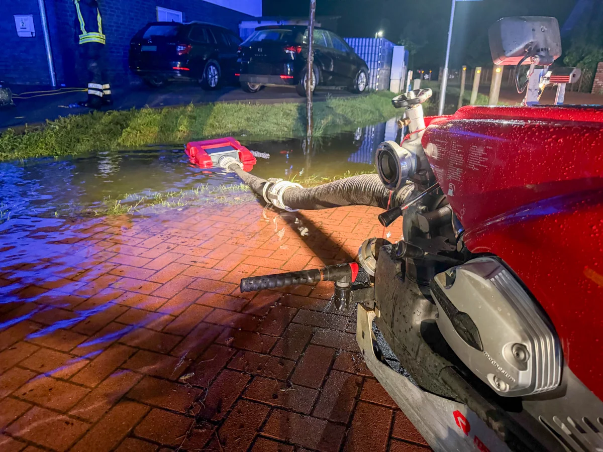 Eine Feuerwehrpumpe ist auf eine überflutete Straße gerichtet, um Wasser abzupumpen. Im Hintergrund sind ein Feuerwehrmann und parkende Fahrzeuge sichtbar.