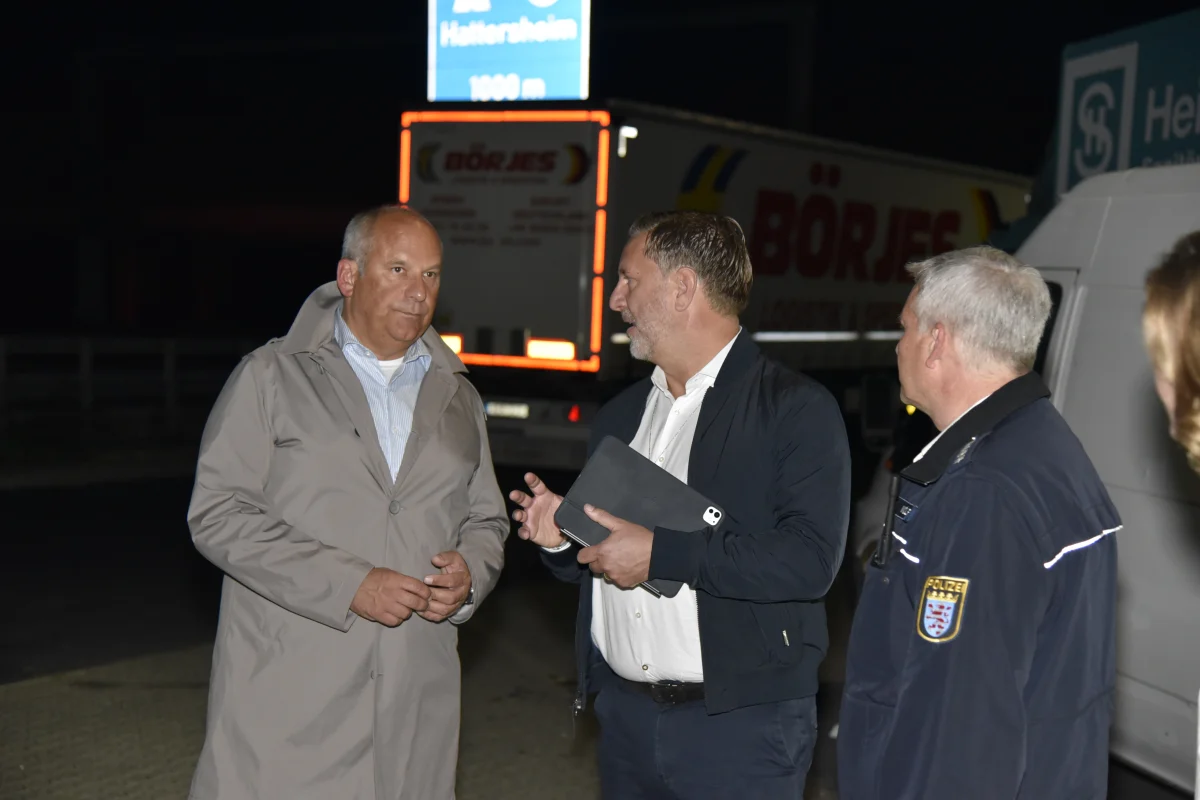 Drei Männer stehen bei Nacht vor einem geparkten LKW und sind im Gespräch vertieft. Einer trägt eine Polizeiuniform.