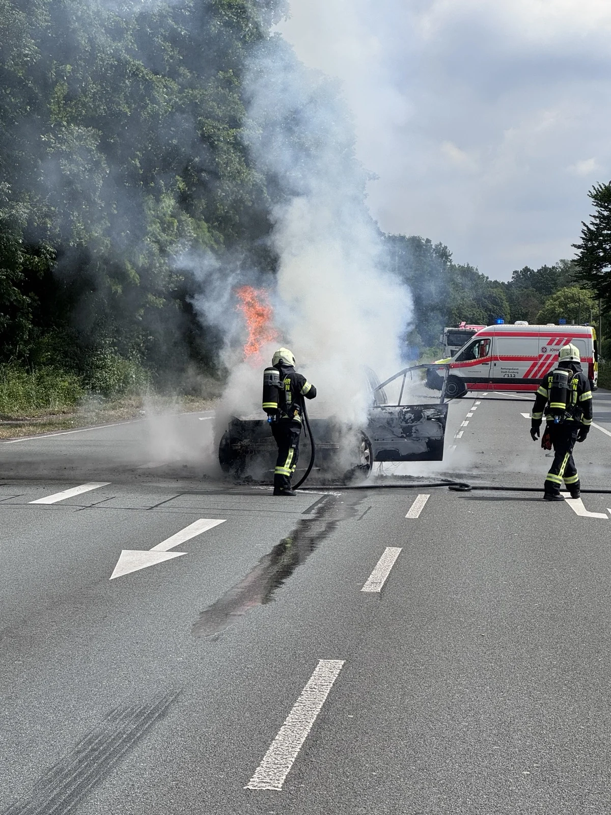 Zwei Feuerwehrleute bekämpfen ein Feuer in einem Auto auf einer Landstraße. Dicker Rauch steigt auf, während ein Rettungswagen im Hintergrund steht.