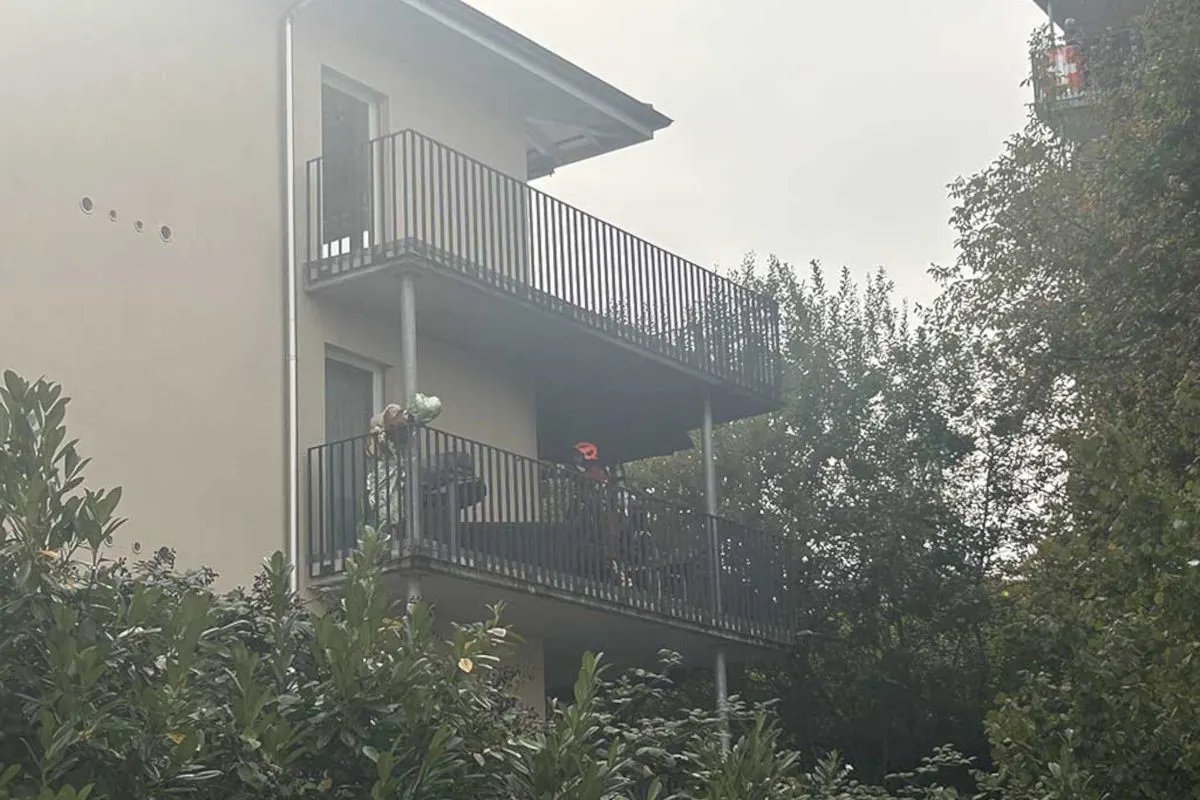 Zwei Personen stehen auf einem Balkon eines Wohnhauses, während Rauch das Gebäude umgibt. Grünpflanzen sind im Vordergrund sichtbar.
