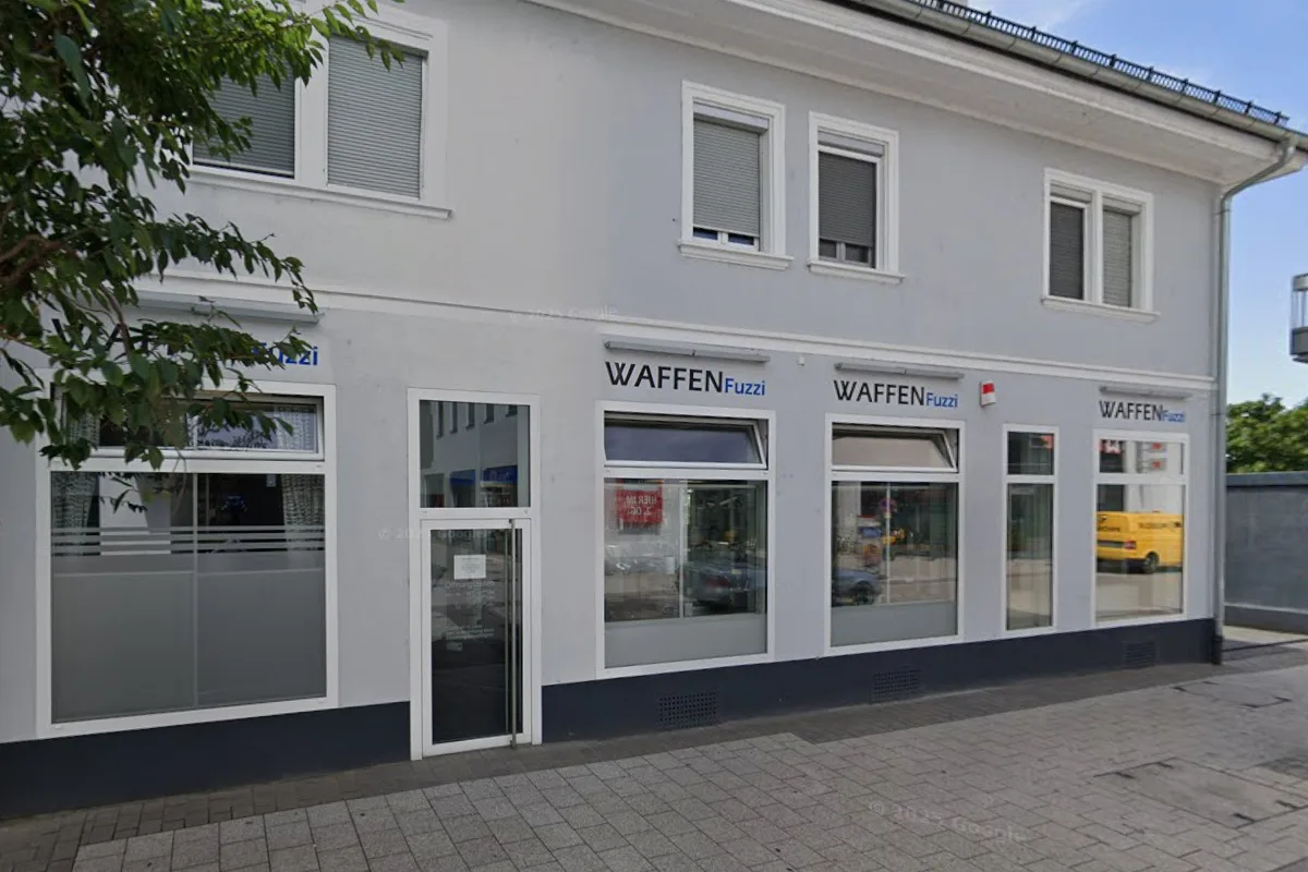 Ein zweistöckiges Gebäude mit weißer Fassade und dem Schriftzug 'WAFFEN Fuzzi' über mehreren Schaufenstern. Vor dem Gebäude ist ein Gehweg zu sehen.