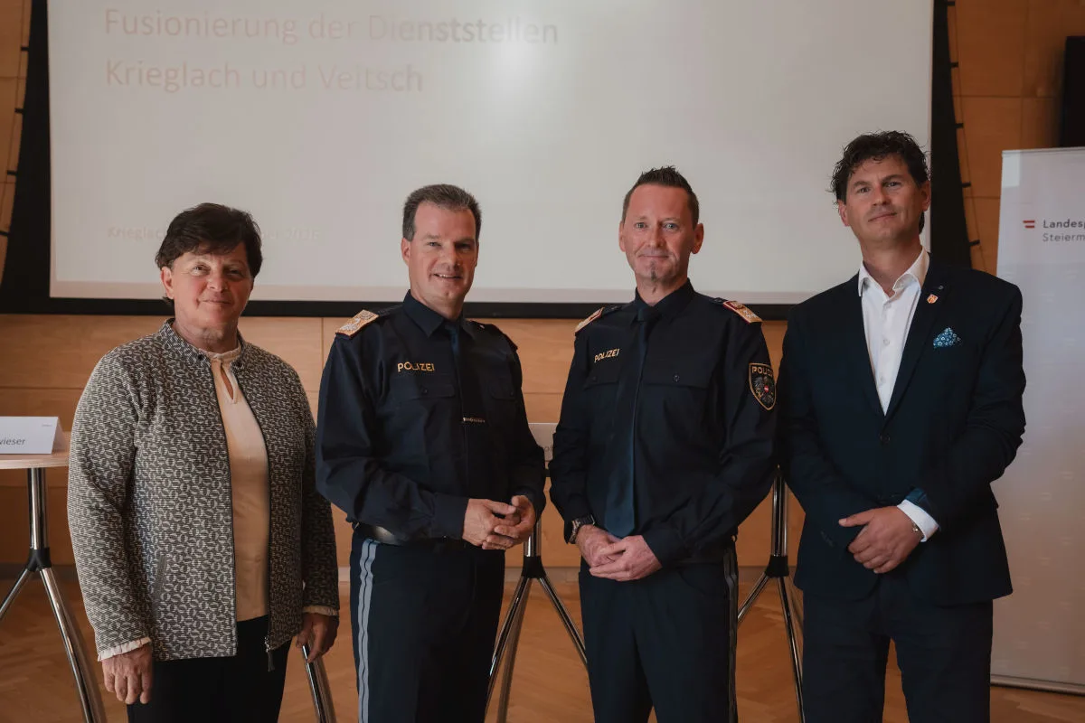 Vier Personen, darunter zwei in Polizeiuniform, stehen nebeneinander vor einer Leinwand mit Präsentation. Ein Schild und Logo sind im Hintergrund sichtbar.