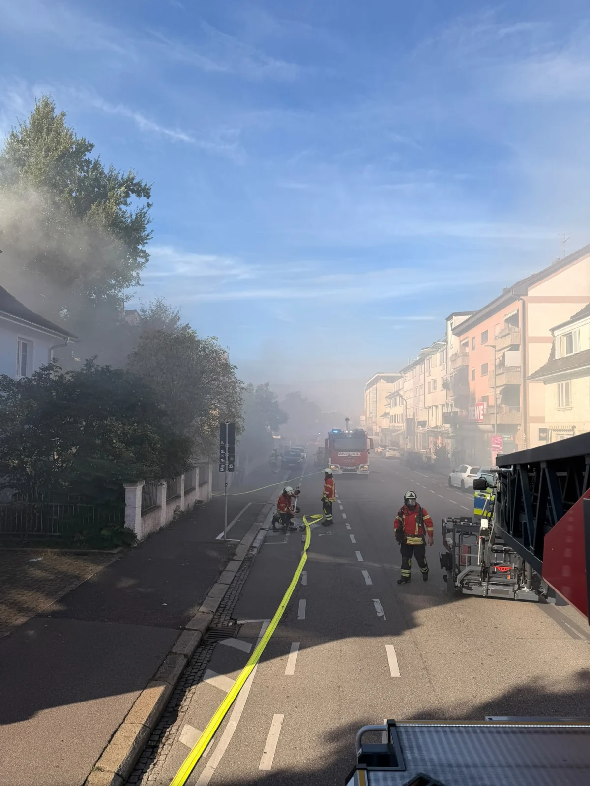Mehrere Feuerwehrleute stehen mit einem Löschschlauch auf einer Straße, während dichter Rauch das Gebiet einhüllt. Im Hintergrund sind Einsatzfahrzeuge zu sehen.