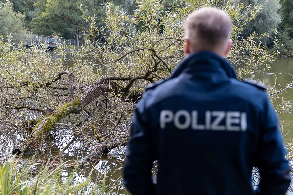 Ein Polizist in dunkler Uniform steht am Ufer eines Gewässers und schaut auf umgestürzte Bäume, die ins Wasser ragen. Im Hintergrund sind dichte, grüne Bäume zu sehen.