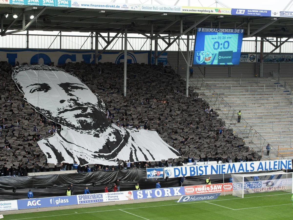 Ein riesiges Banner mit einem Gesicht wird von Fans in einem vollbesetzten Fußballstadion hochgehalten. Im Hintergrund ist ein Display mit Spielinformationen zu sehen.