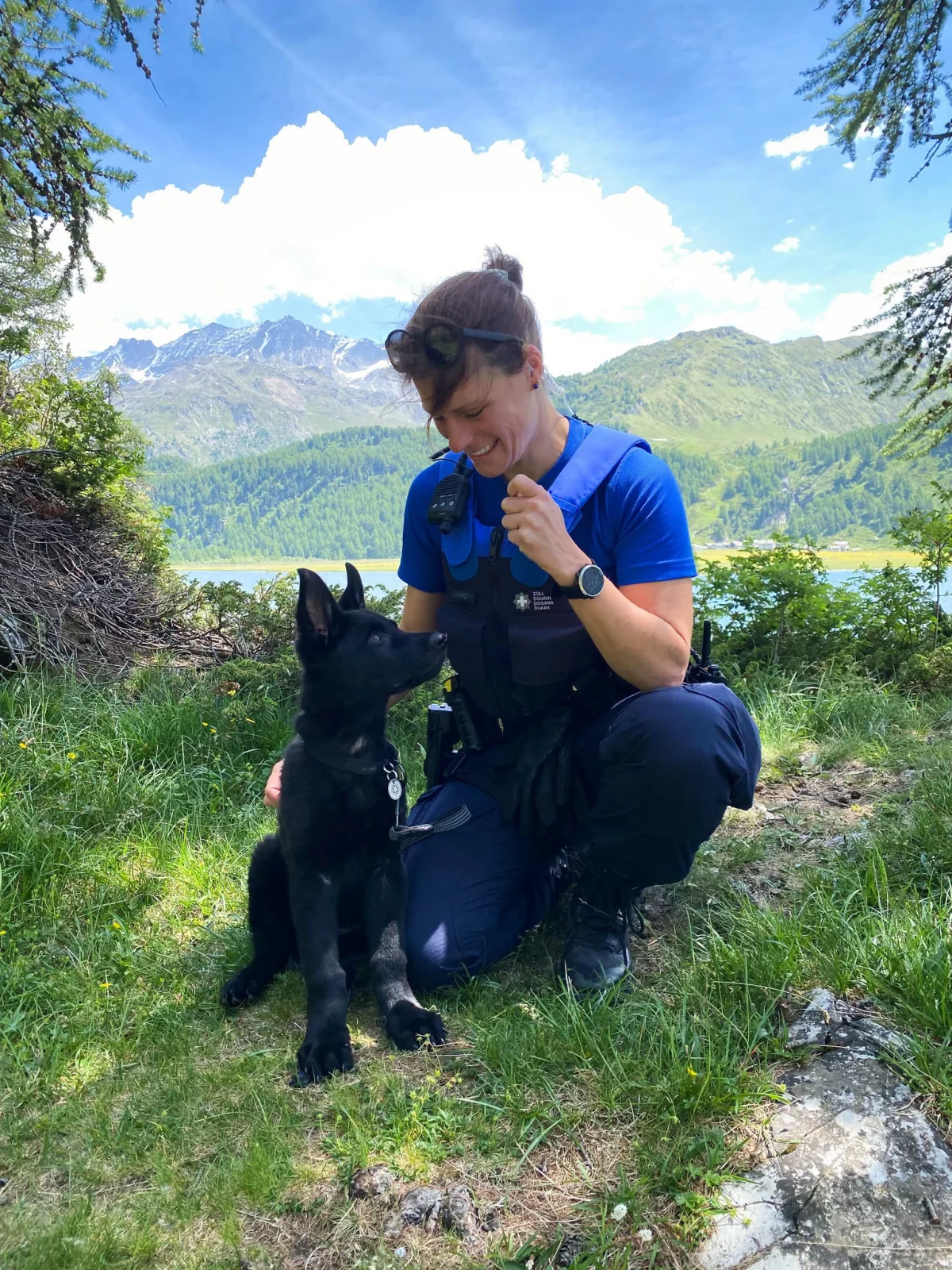 Eine Frau in Polizeiuniform interagiert mit einem schwarzen Hund vor einer alpinen Bergkulisse mit blauem Himmel.