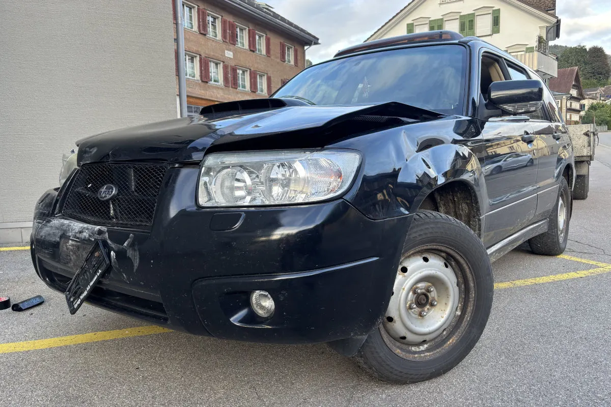 Ein schwarzer SUV zeigt Schäden an der Frontpartie und steht schräg in einem Parkplatzbereich. Im Hintergrund sind Wohnhäuser sichtbar.