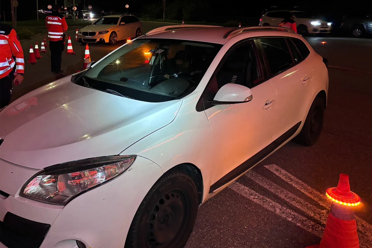 Zwei Polizisten in leuchtenden Westen führen nachts eine Verkehrskontrolle mit einem weißen Auto durch. Der Bereich ist mit Kegeln abgesichert.