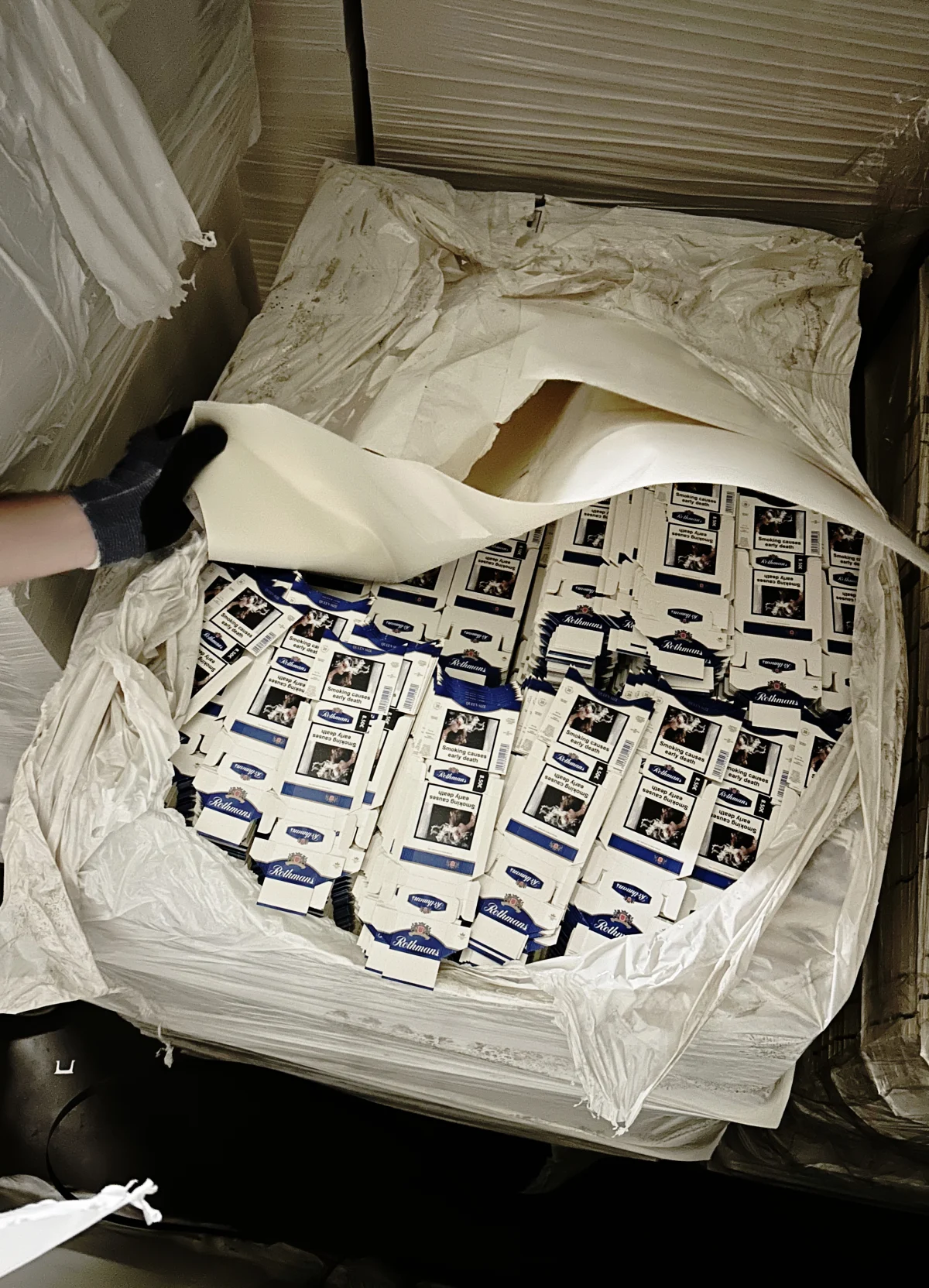 Eine behandschuhte Hand entfernt Plastikfolie von Kartons, die mit Zigarettenpackungen gefüllt sind. Die Zigarettenpackungen liegen dicht gestapelt.