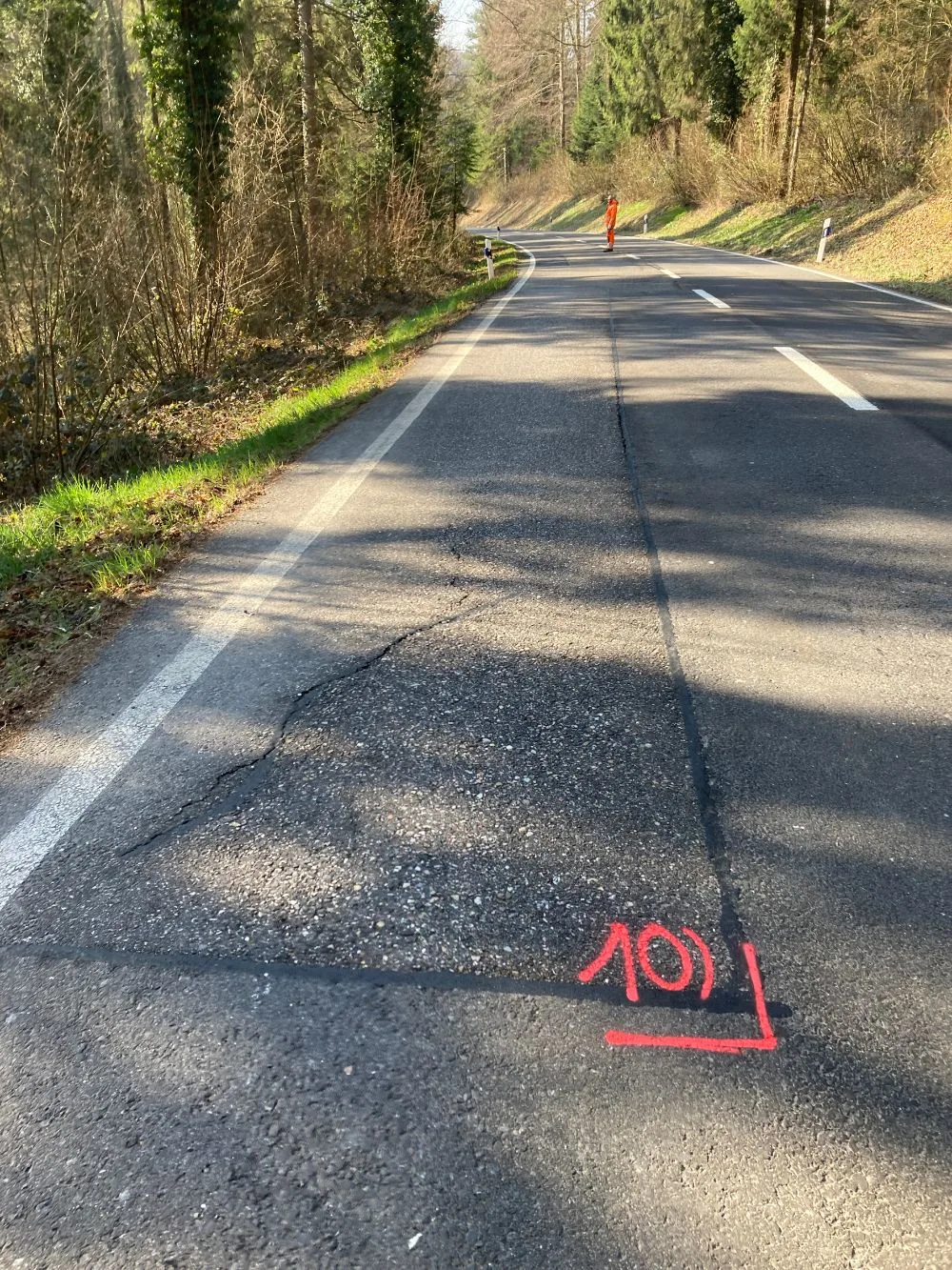 Eine Landstraße mit Rissen und einer roten Markierung ist sichtbar, ein Arbeiter in orangener Warnkleidung steht in der Ferne.