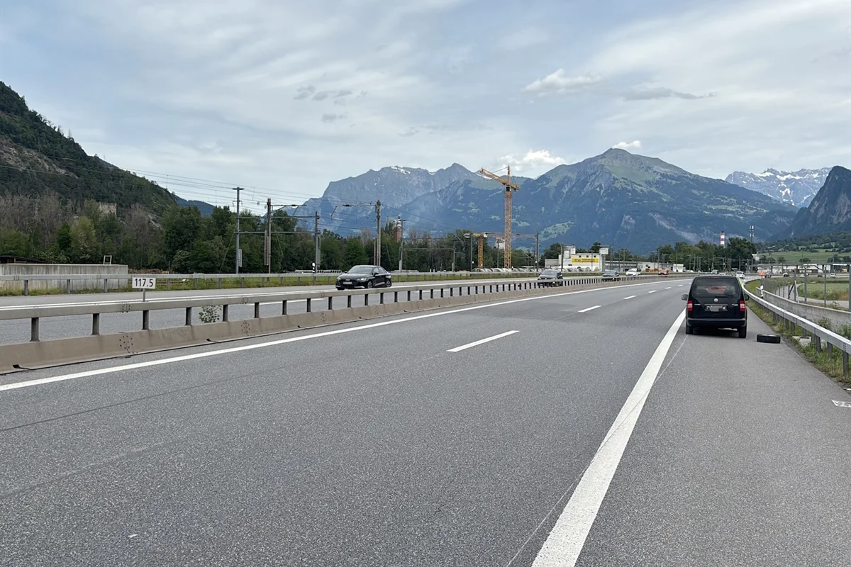 Ein Auto steht am rechten Rand eines Highways, mit einem abmontierten Reifen daneben. Im Hintergrund sind Berge sichtbar.