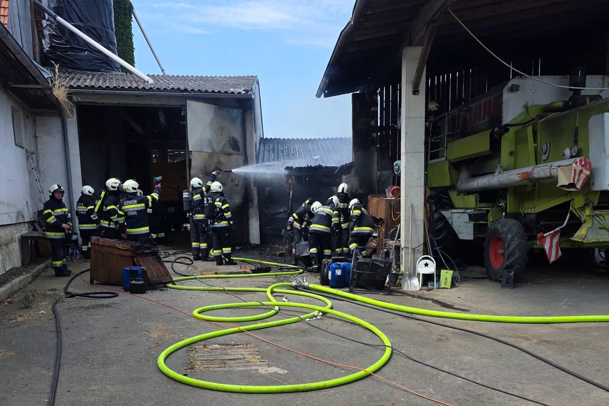 Mehrere Feuerwehrleute in Schutzkleidung löschen ein Feuer mit Wasserschläuchen in einer Scheune. Im Vordergrund sind grüne Schläuche zu sehen.