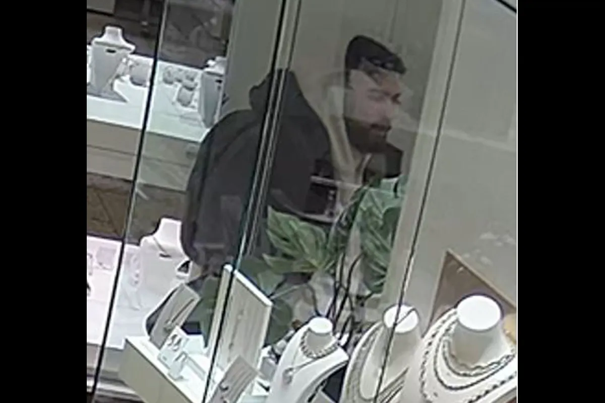 Ein unidentifizierter Mann wird durch ein Schaufenster in einem Juweliergeschäft beobachtet. Er trägt einen dunklen Hoodie und steht neben Schmuckauslagen.
