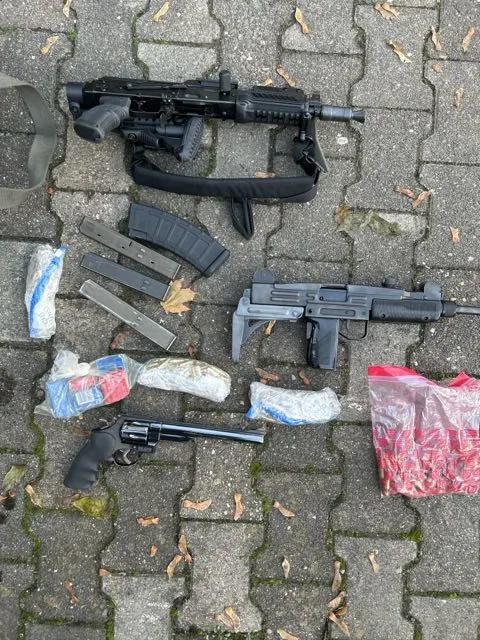 Ein Sturmgewehr, eine Maschinenpistole, ein Revolver und mehrere Magazine liegen auf einem Pflastersteinboden zusammen mit Plastiktüten.