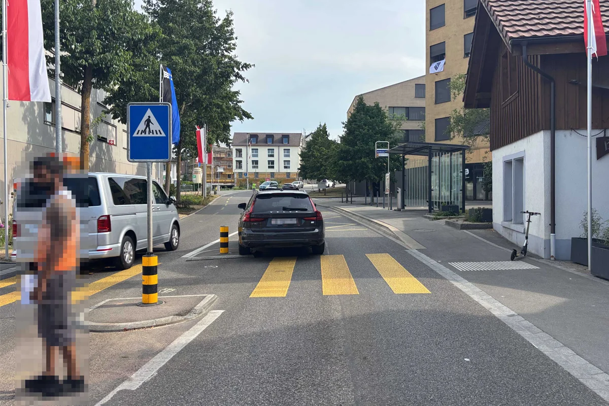 Ein schwarzes Auto ist auf einem gelben Zebrastreifen in einer städtischen Straße geparkt. Im Hintergrund sind Gebäude und Bäume sichtbar.