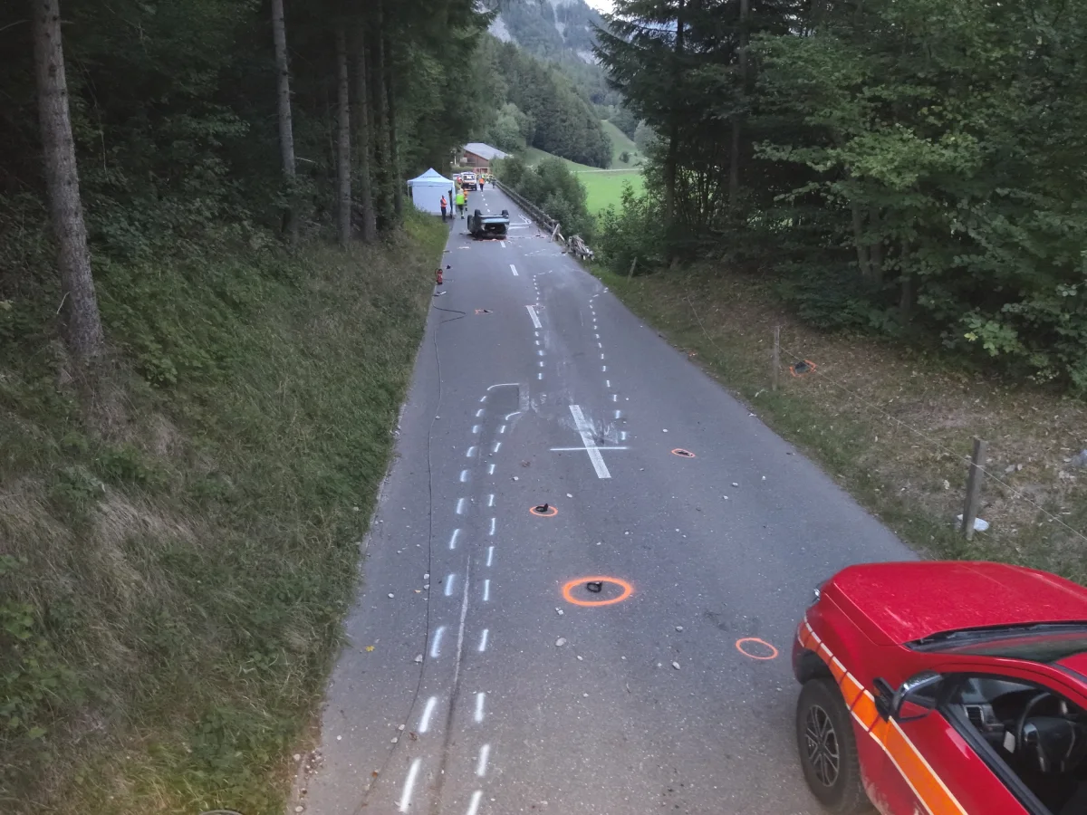 Eine Landstraße mit Markierungen, die einen Unfall skizzieren, ein rotes Fahrzeug steht am Straßenrand. Die Straße ist von Bäumen umgeben.